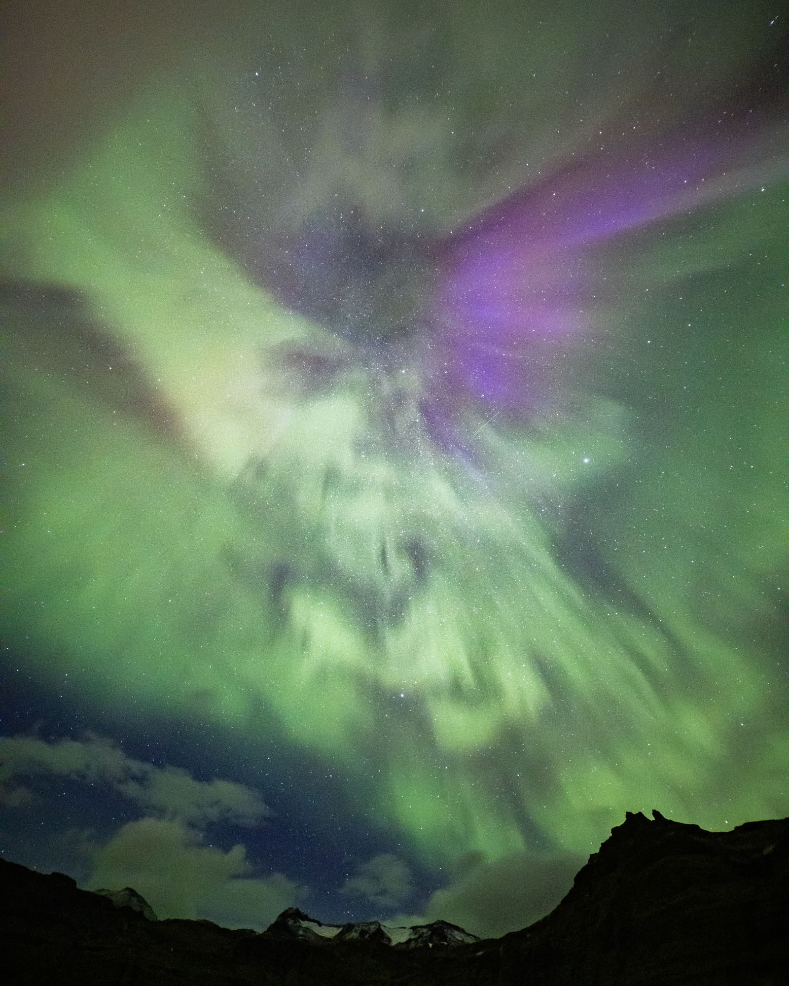 A swirling green and purple aurora forming wing-like shapes across the sky above a silhouetted Icelandic mountain.