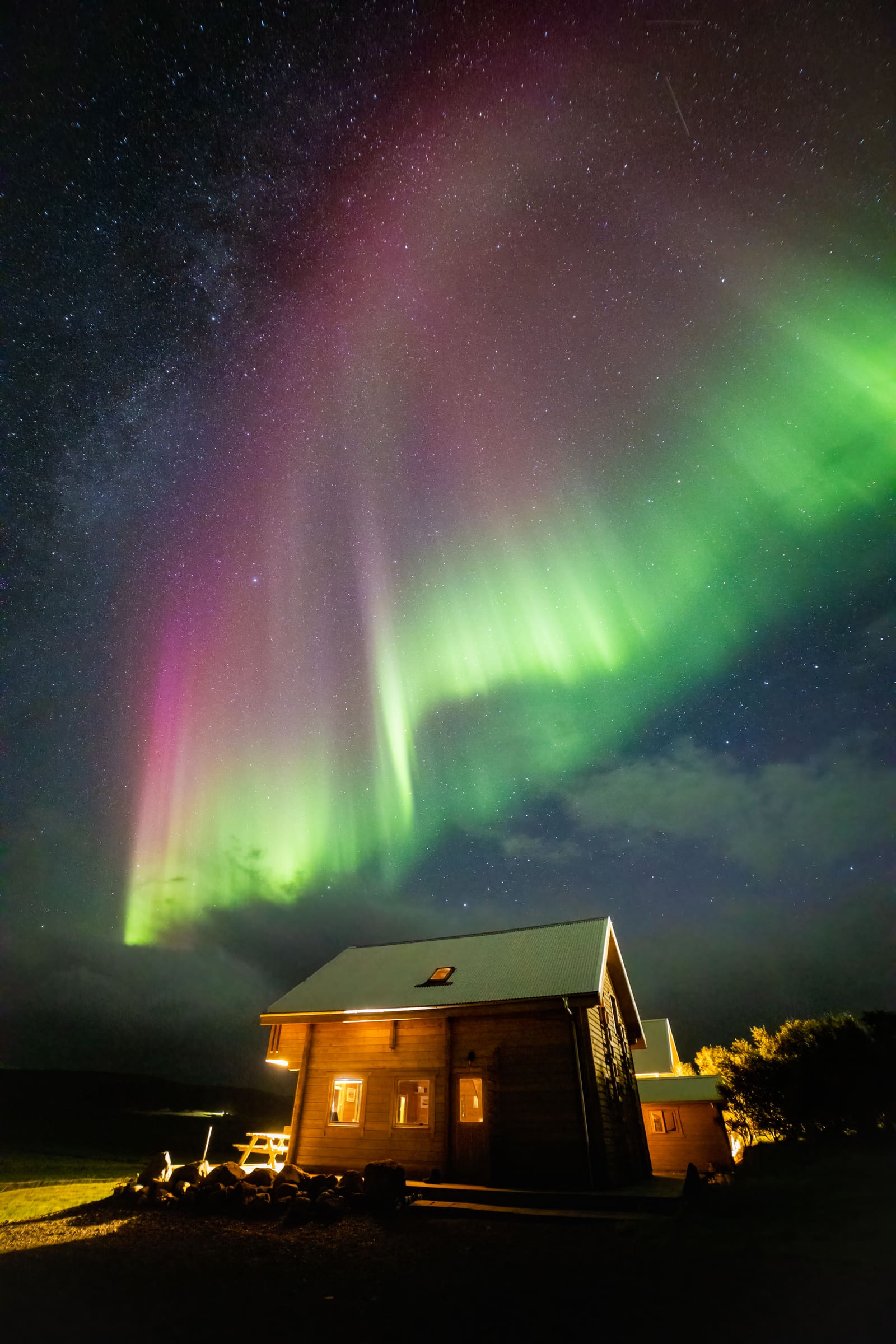 Tall pink and green aurora pillars rising above a lit wooden cabin with a woodpile in the foreground, at Reykholar, Iceland.