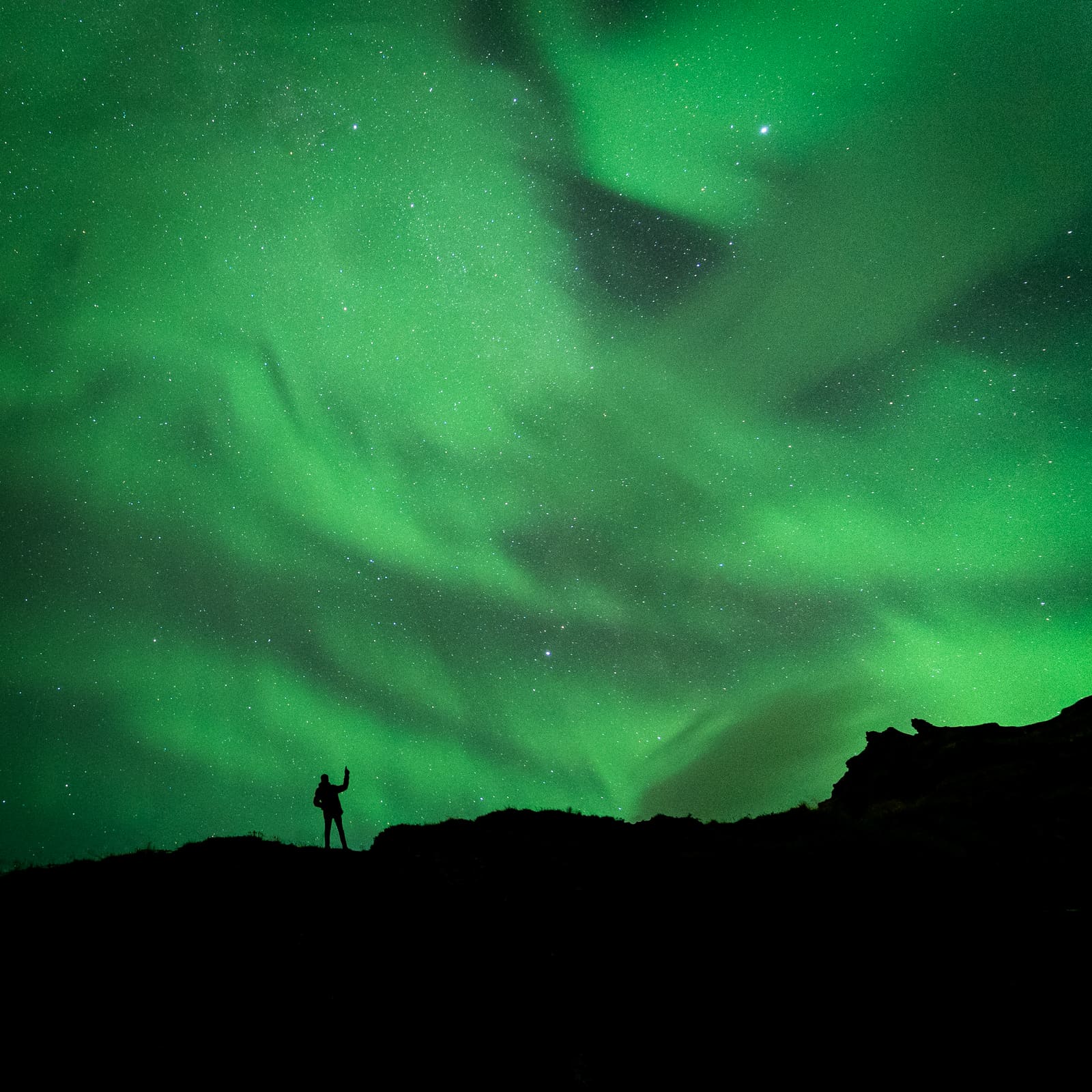 A person silhouetted beneath a bright green aurora and starry sky in Iceland.