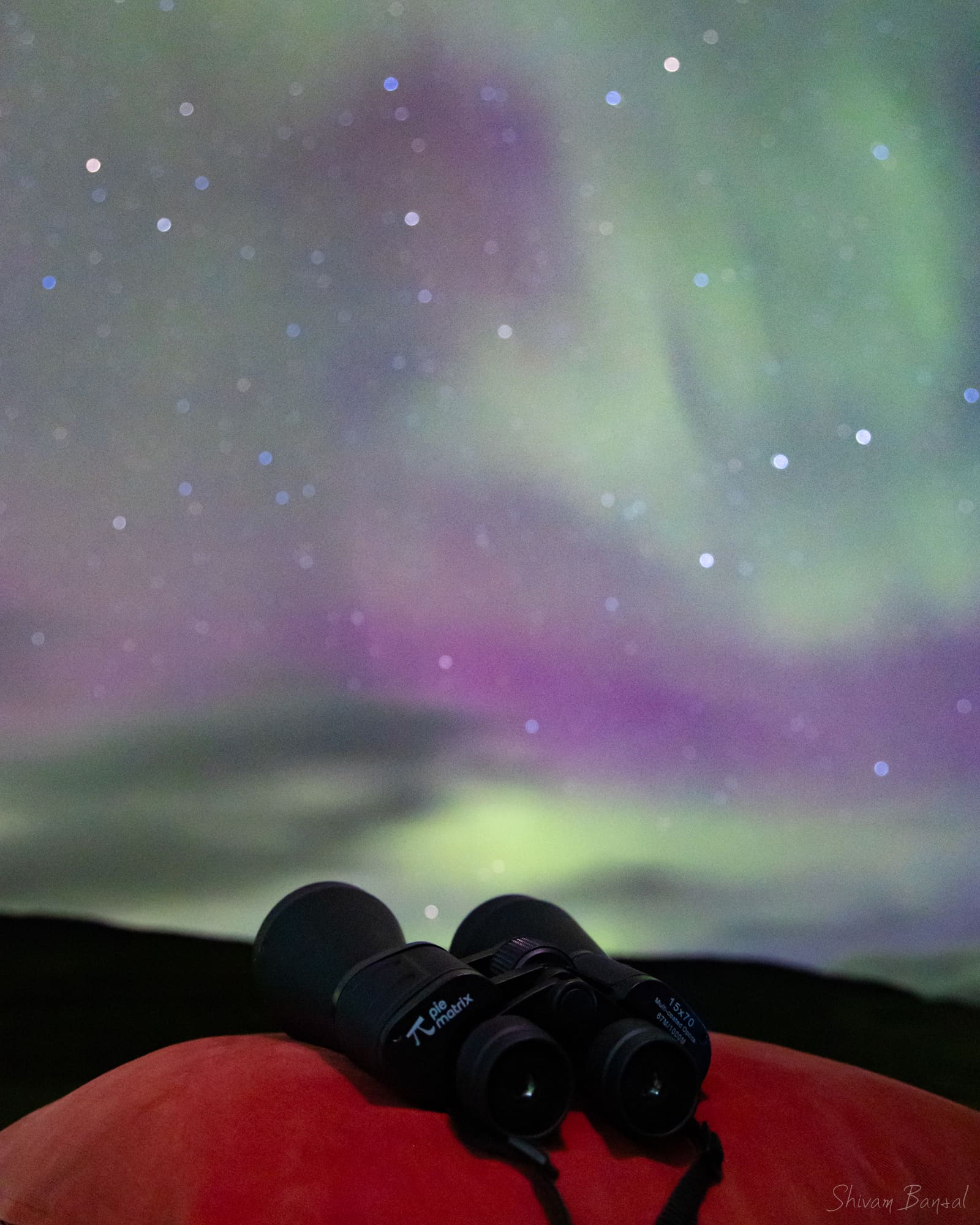 Binoculars in the foreground beneath a colorful pink, purple, and green aurora and starry sky.