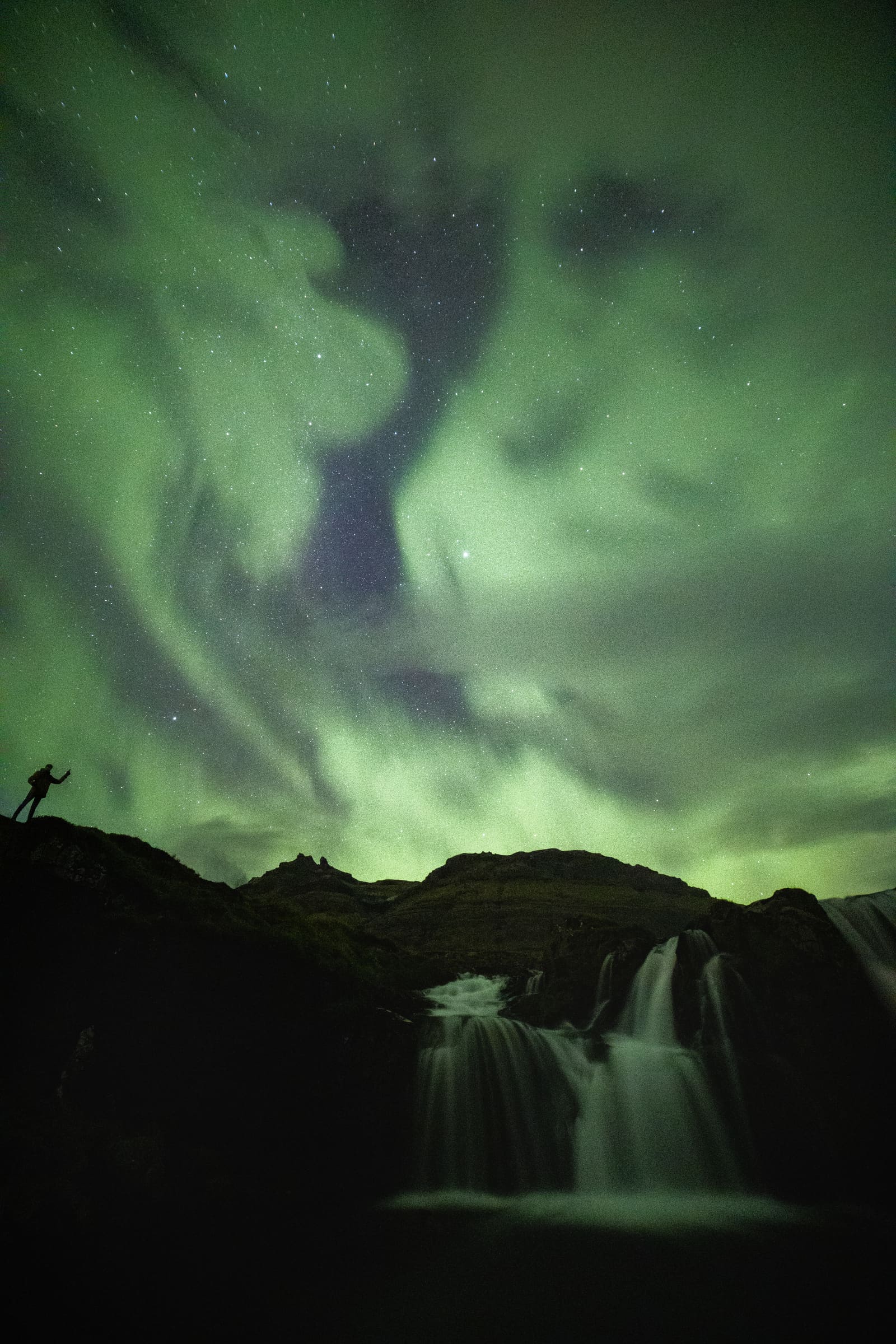 Green aurora bands over a waterfall in Iceland, with a person silhouetted on the ridge to the left.
