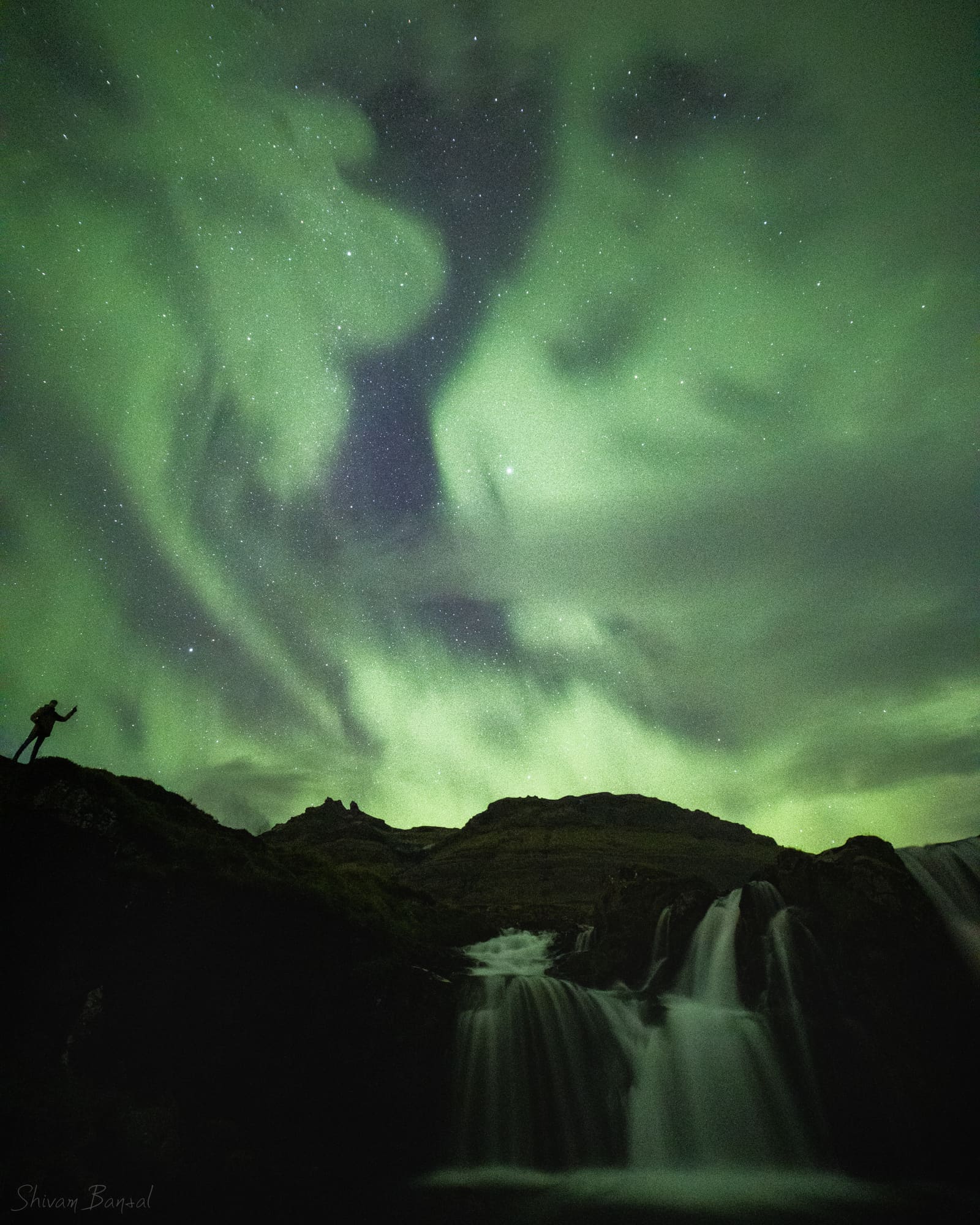 A swirling green aurora overhead, with a small silhouetted figure on a ridge above a waterfall.