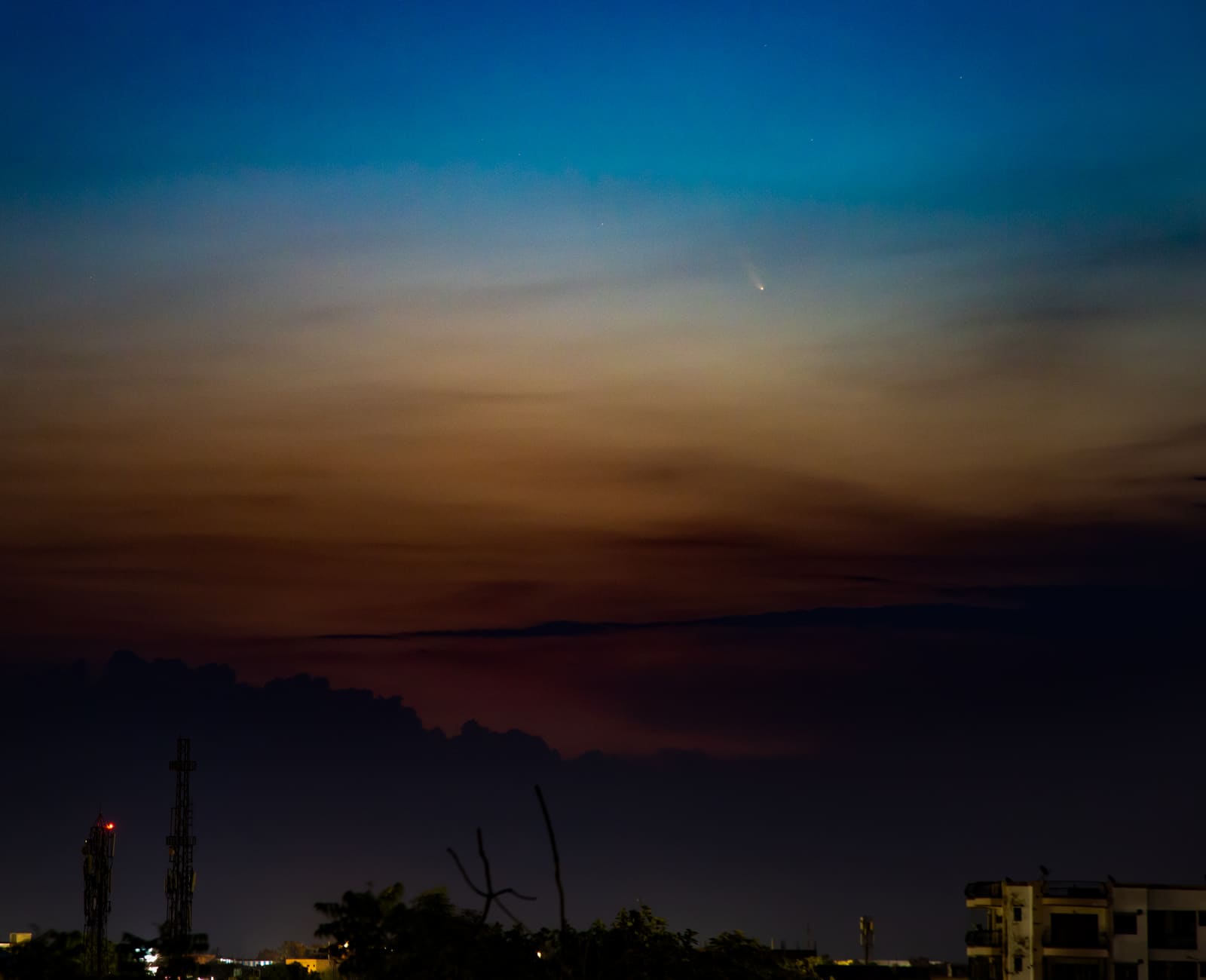 Wide shot of Comet NEOWISE low in an orange-to-blue twilight sky above a city skyline with dark building and crane silhouettes.