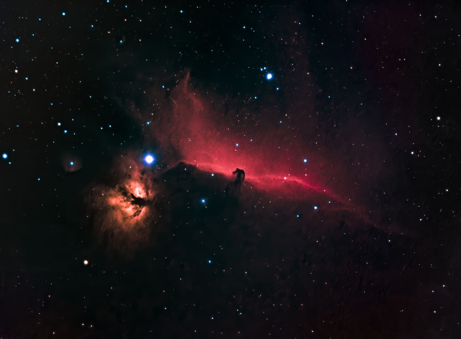 A dark horse-head-shaped nebula against red gas clouds, with a bright star and an orange flame-like nebula to the left.