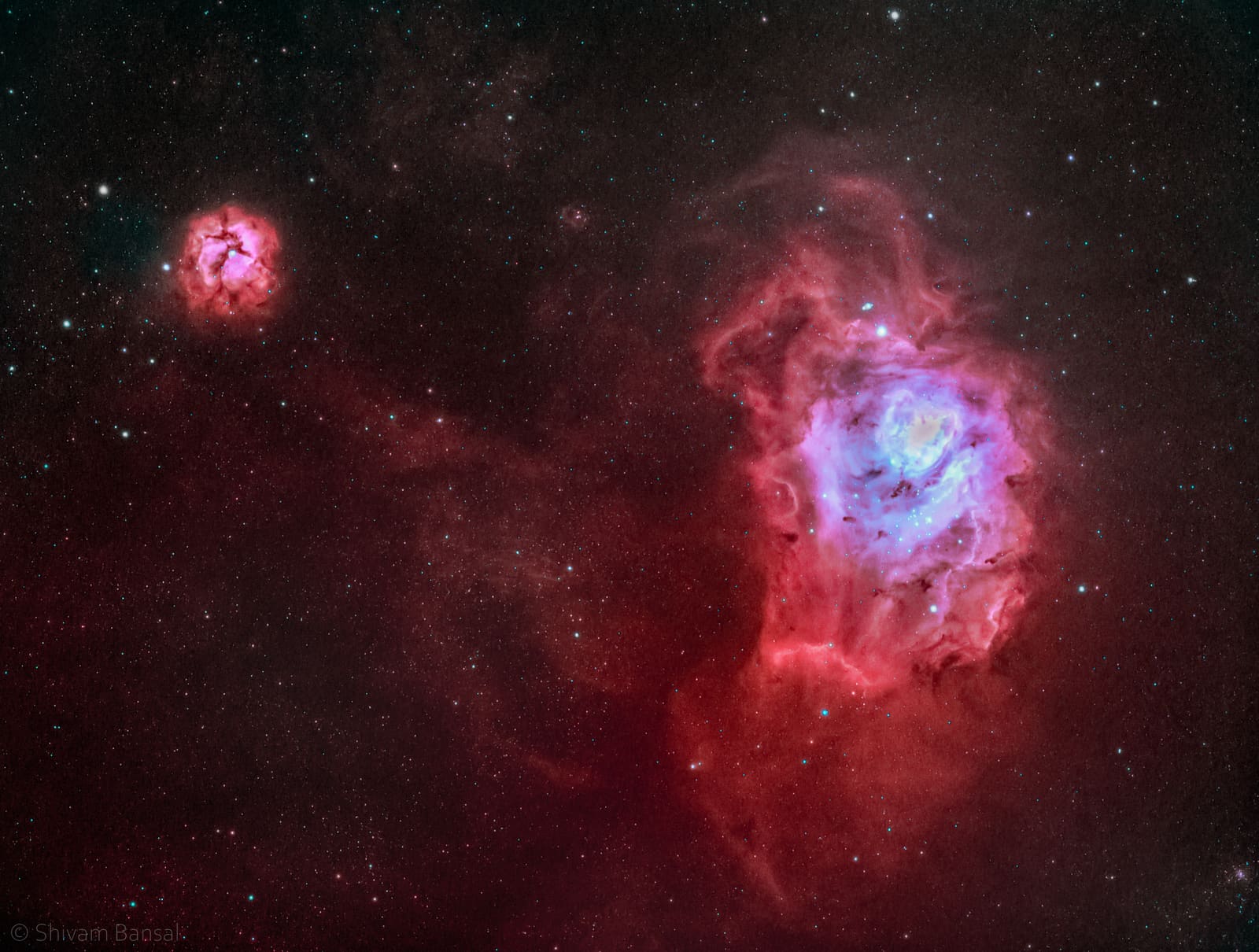 A large pink emission nebula with dark dust lanes threading through a bright core, and a smaller separate circular pink nebula in the upper left corner.