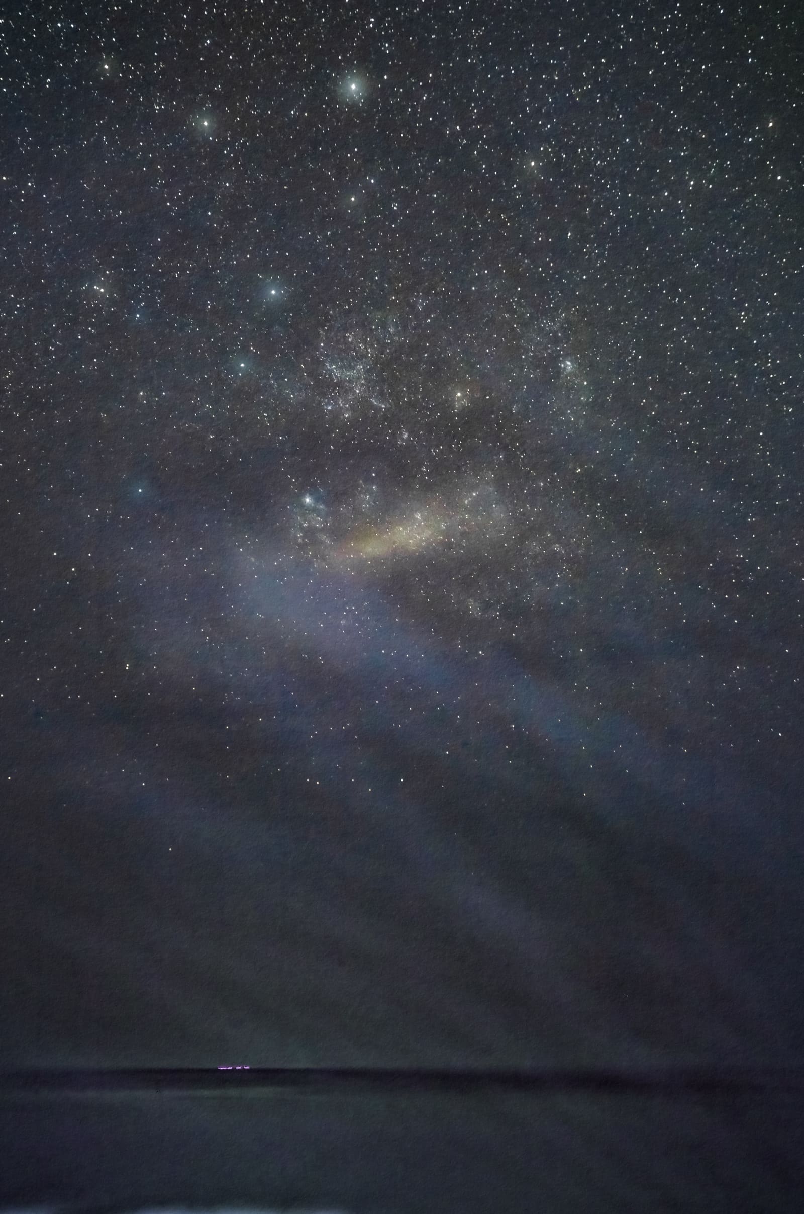 A wide night sky scene with a faint hazy patch of light low above a dark sea horizon, surrounded by stars.