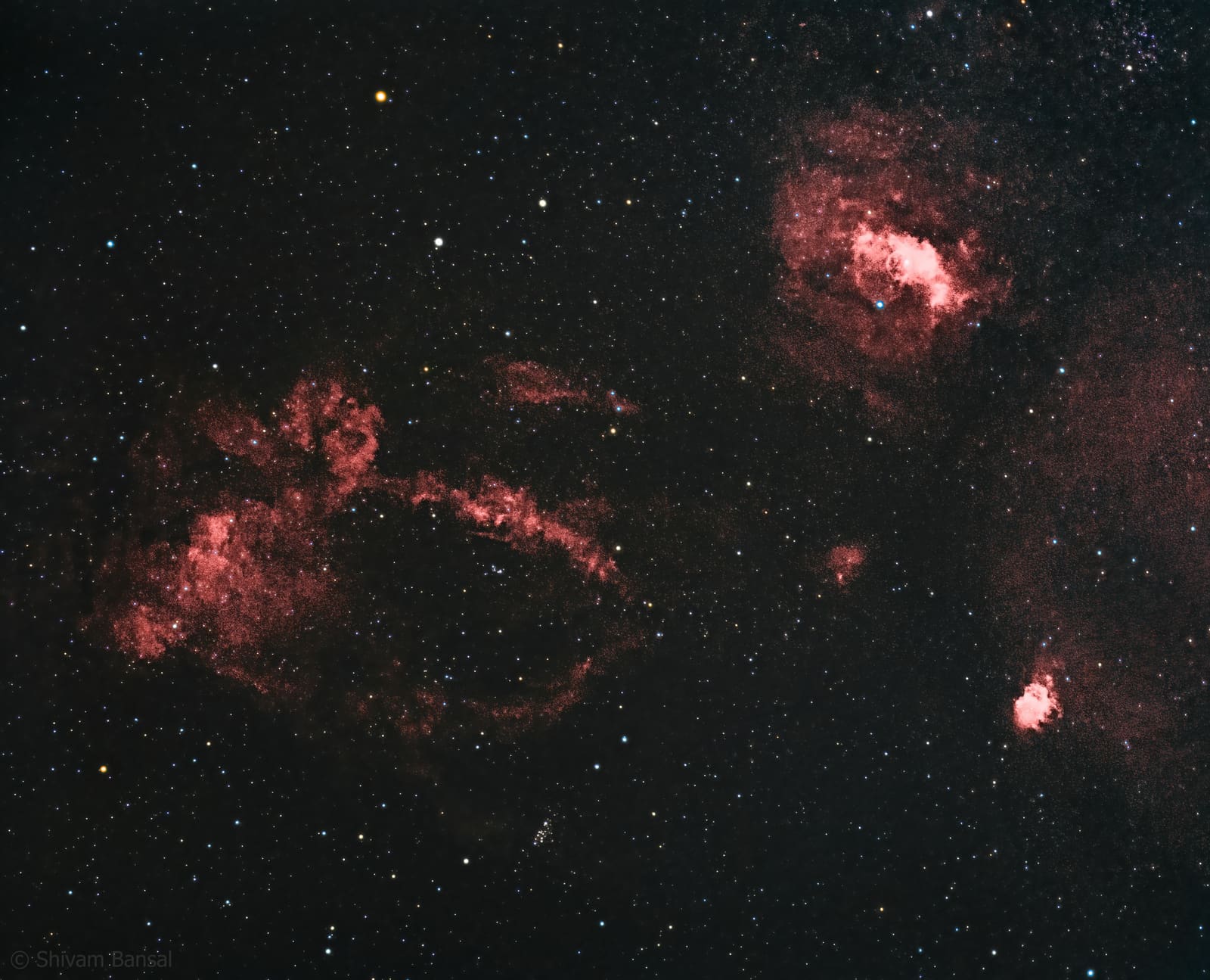 Two separate patches of red nebulosity, a larger claw-like looping structure and a smaller rounder nebula, set in a starfield.