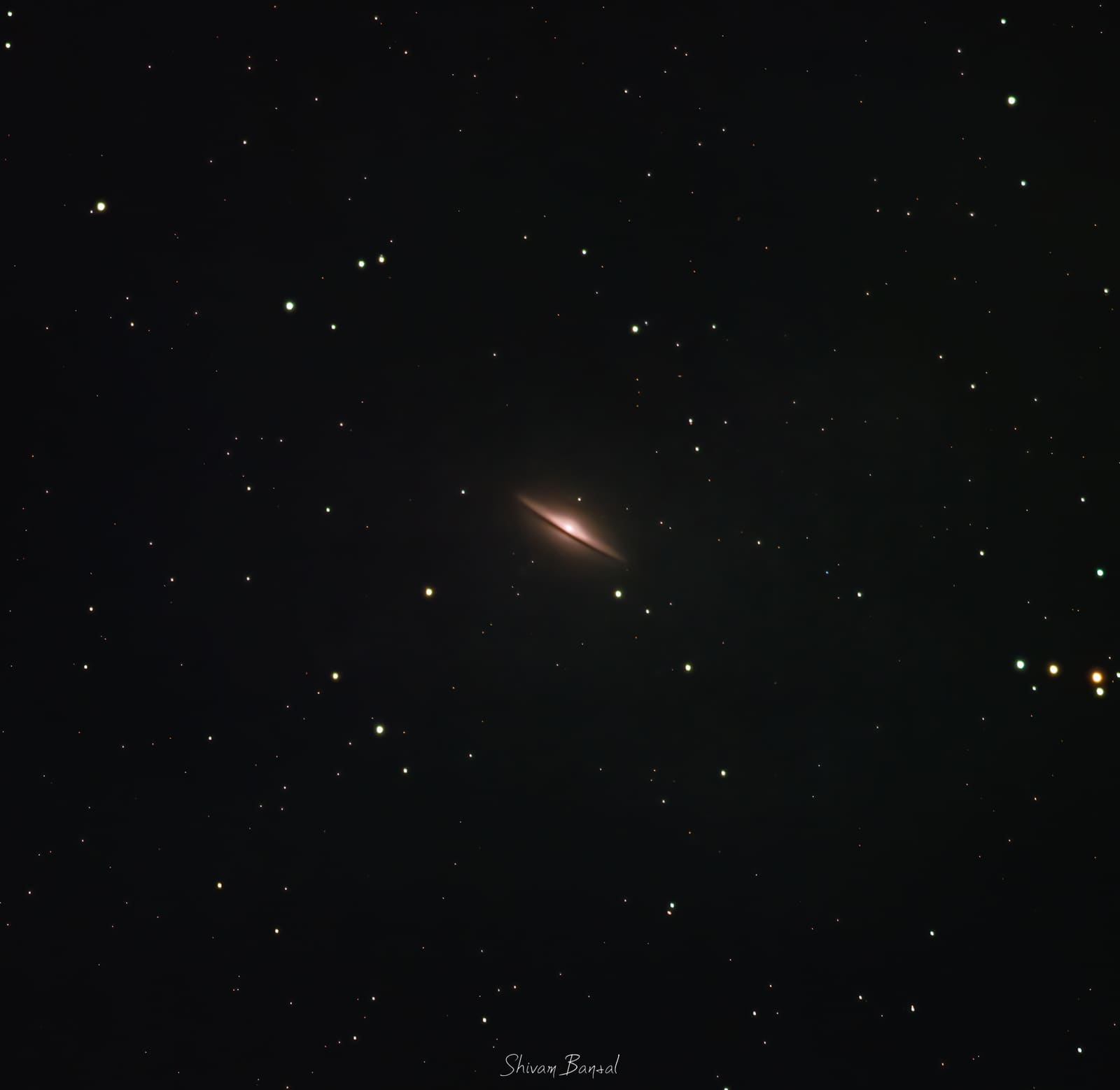 A small, thin, edge-on galaxy with a bright central bulge and a dark dust lane, surrounded by scattered stars.