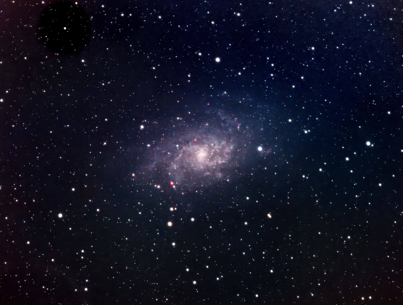 A face-on spiral galaxy with loosely wound arms and small pink nebulous knots, set against a starfield.