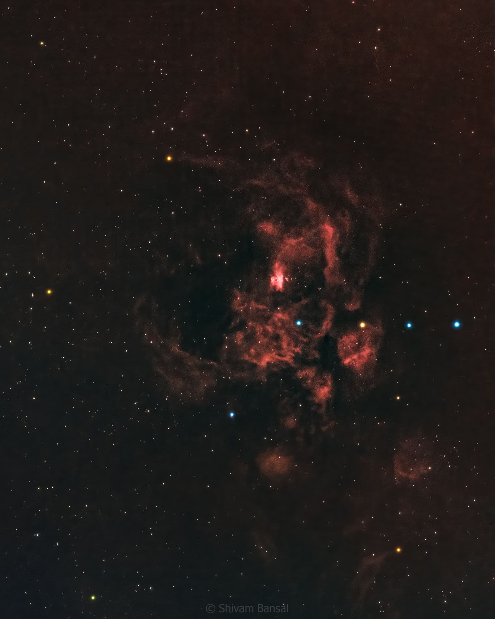 A dim, irregular patch of orange-red nebulosity with a few bright blue stars scattered across a dark starfield.