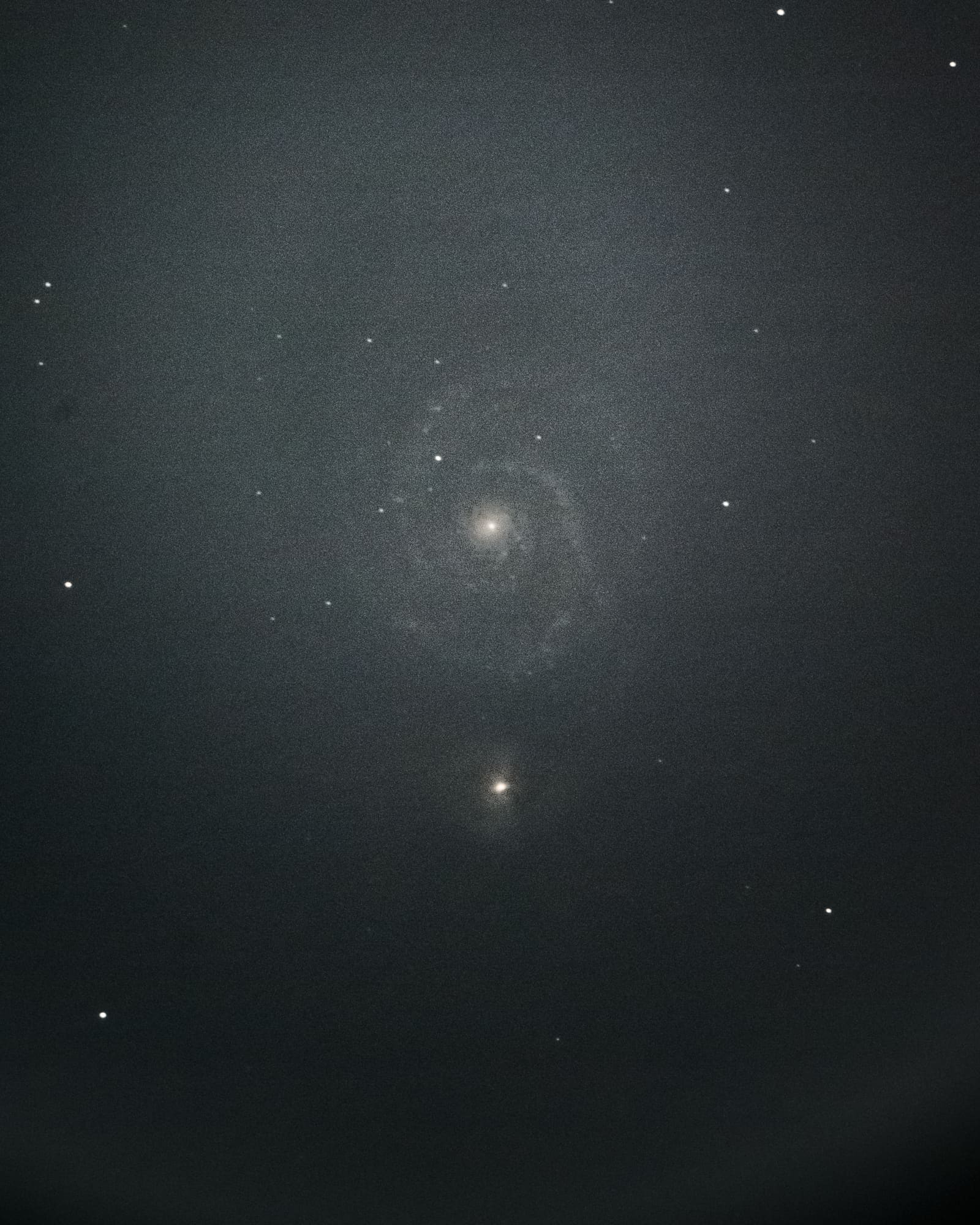 A faint spiral galaxy connected to a smaller, brighter companion galaxy, rendered in muted gray tones against a dark sky.