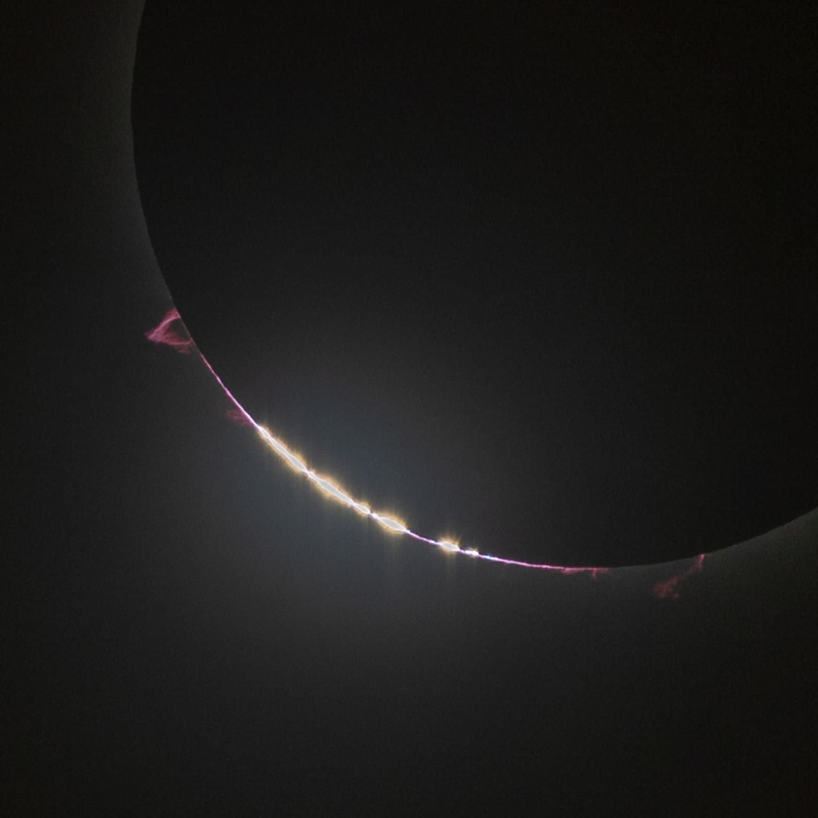 A thin arc of bright beads of light along the edge of a mostly covered sun, with a faint pink glow visible at the rim.