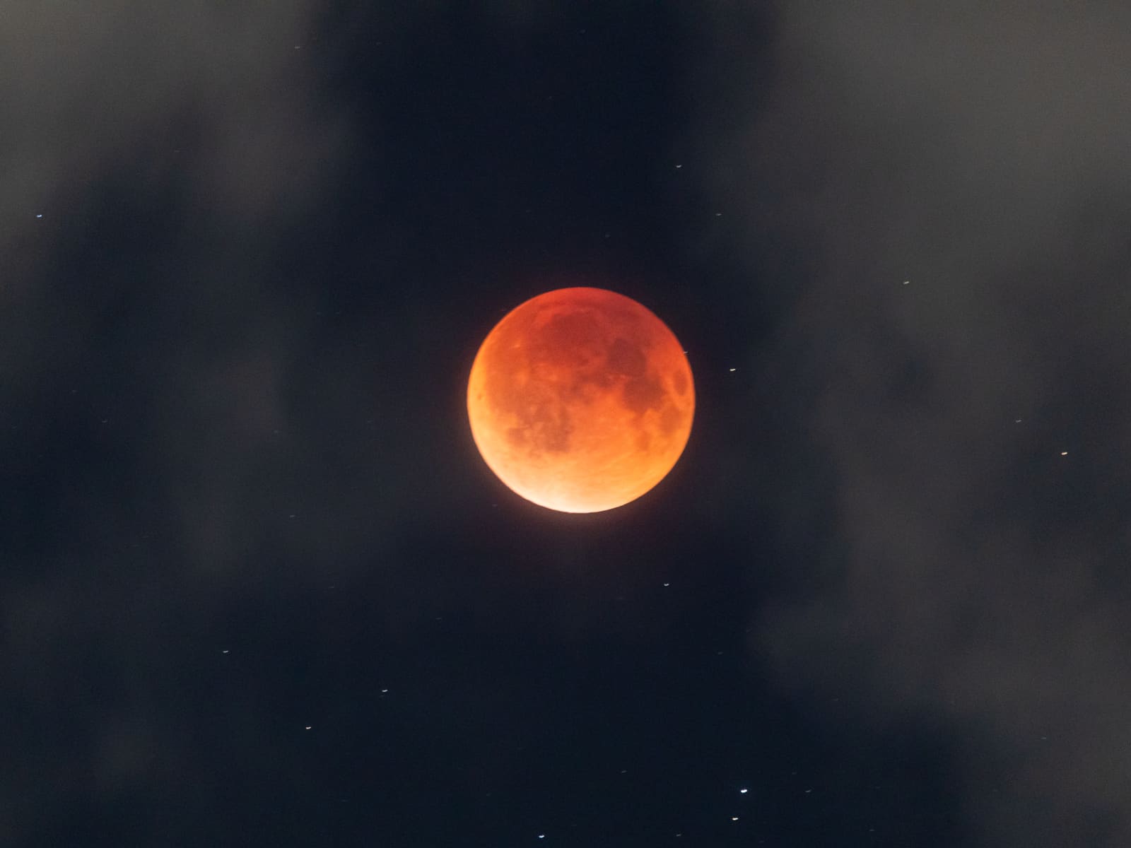 A small red eclipsed moon glowing softly behind thin drifting clouds against a dark sky.
