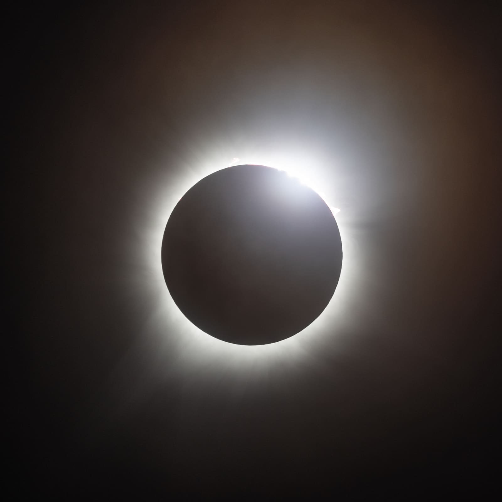 A dark eclipsed sun with a single bright flare of light on its edge, corona faintly visible around the disc.