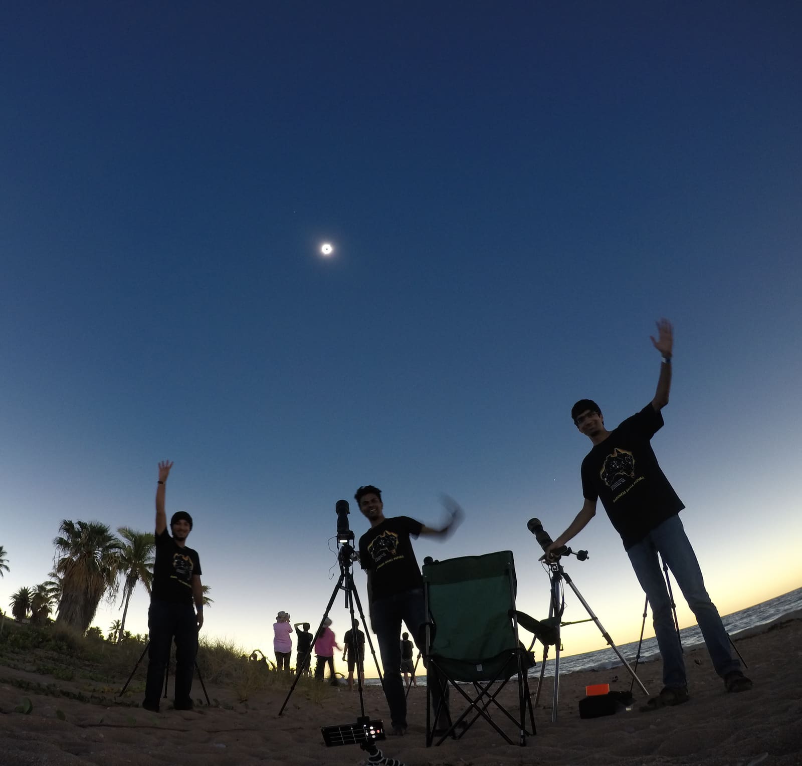 A group of people with telescopes and cameras on tripods standing outdoors under a darkening sky, one person pointing upward.