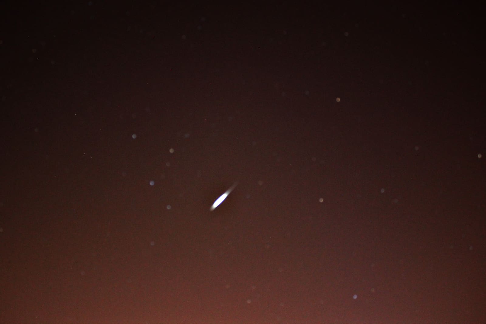 Bright elongated streak of light, wider at the center and tapering at both ends, crossing a dark reddish starry sky, a captured satellite flare.