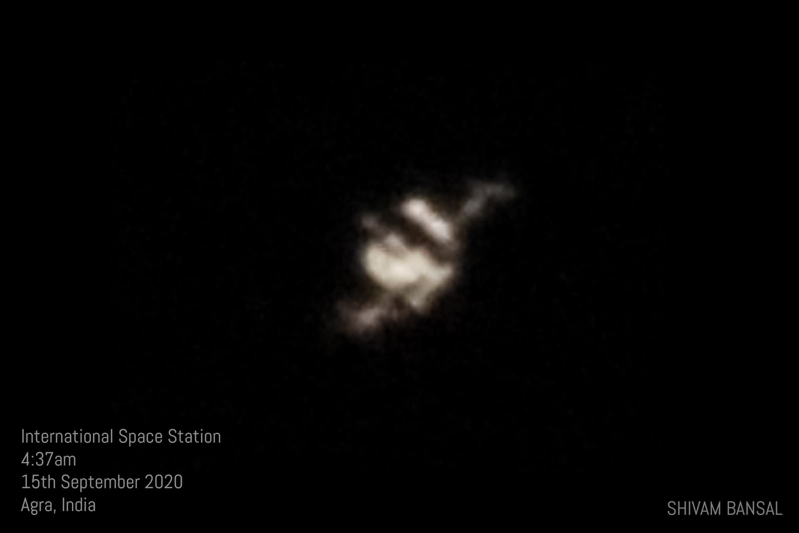 Faint blurry silhouette of the ISS against a black sky, with a text caption giving the time and location of the pre-dawn pass.