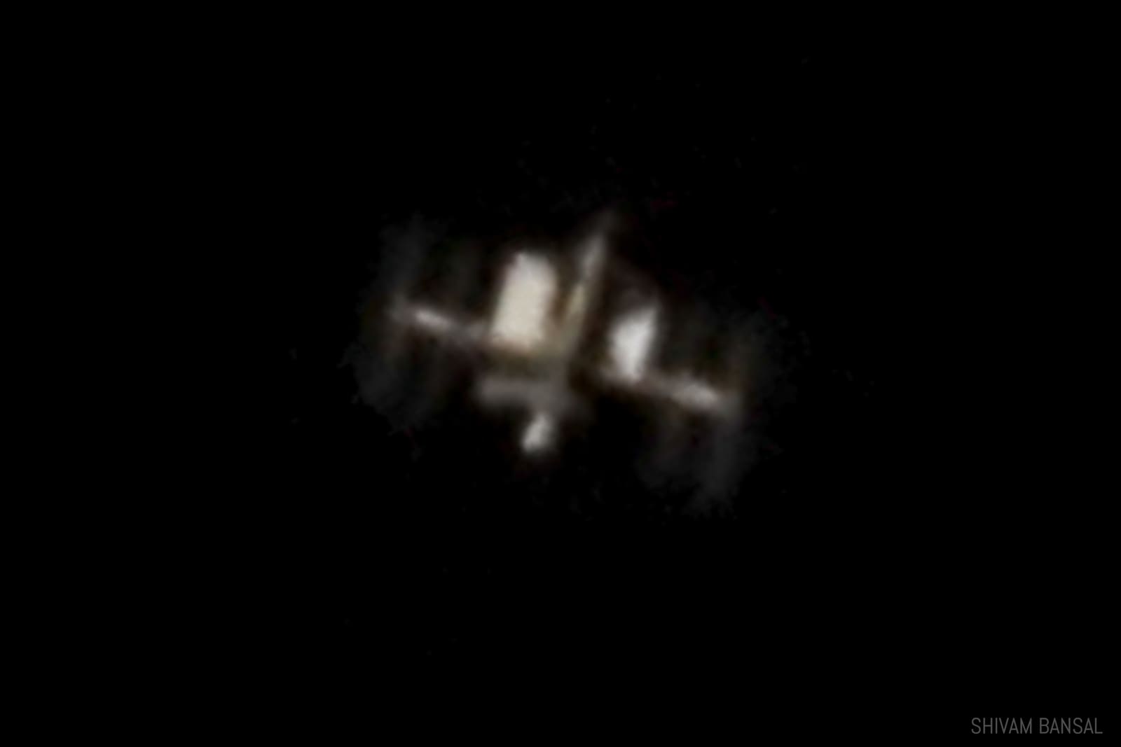 Small but sharp telescopic photo of the International Space Station showing its truss structure and solar panel wings, isolated on black sky.