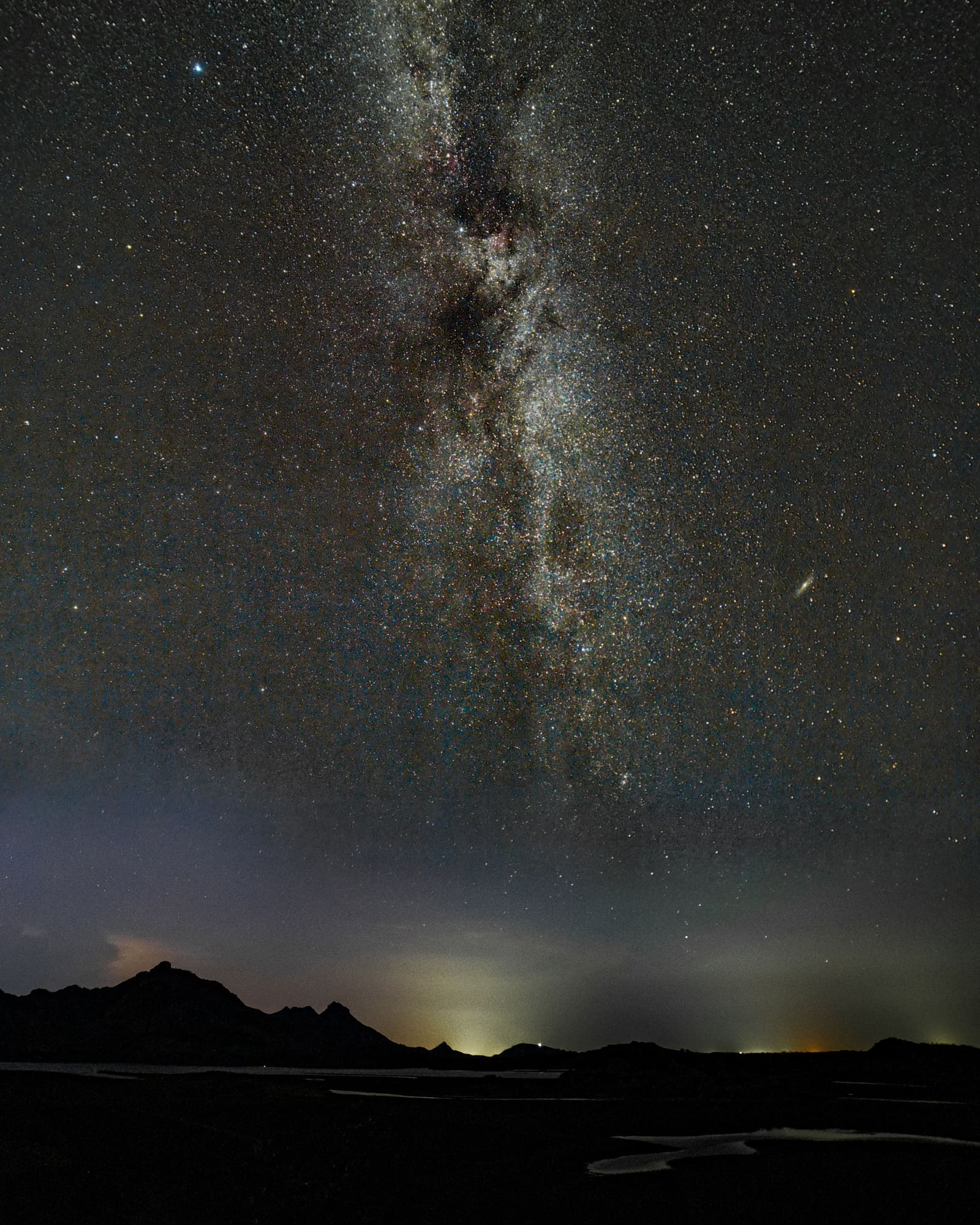 The fainter northern arm of the Milky Way rising over silhouetted hills at Bera, Rajasthan, with a pale glow and distant lights along the horizon.
