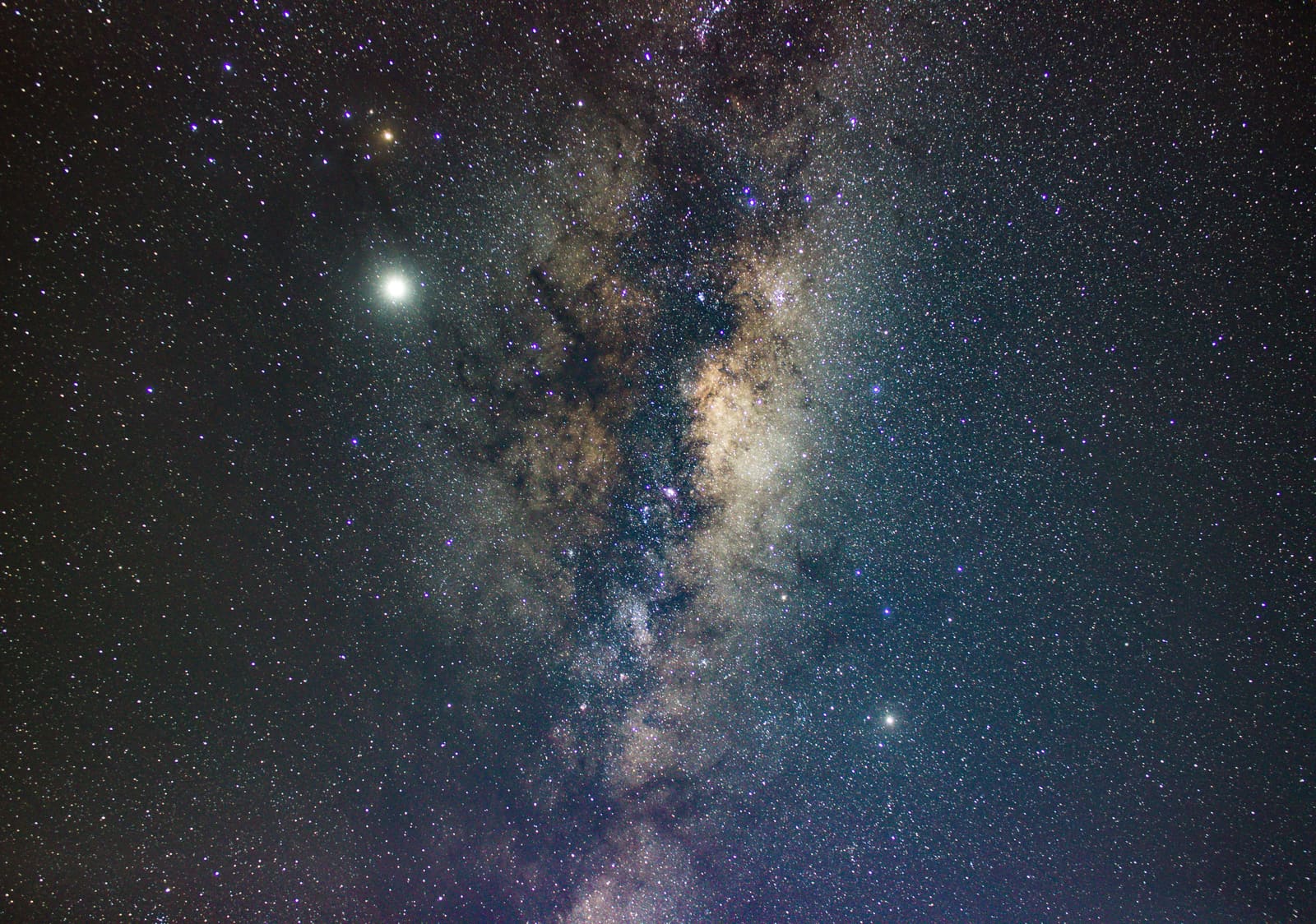 A close view of the Milky Way's core and dust lanes with a very bright planet shining beside it against a dense starfield.