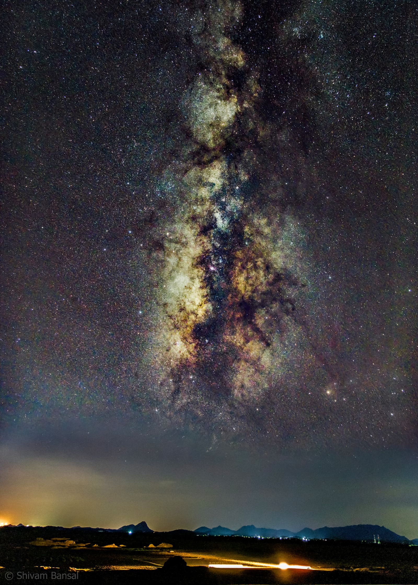 Vertical view of the bright Milky Way core rising above dark silhouetted hills and distant village lights in Jawai, Rajasthan.