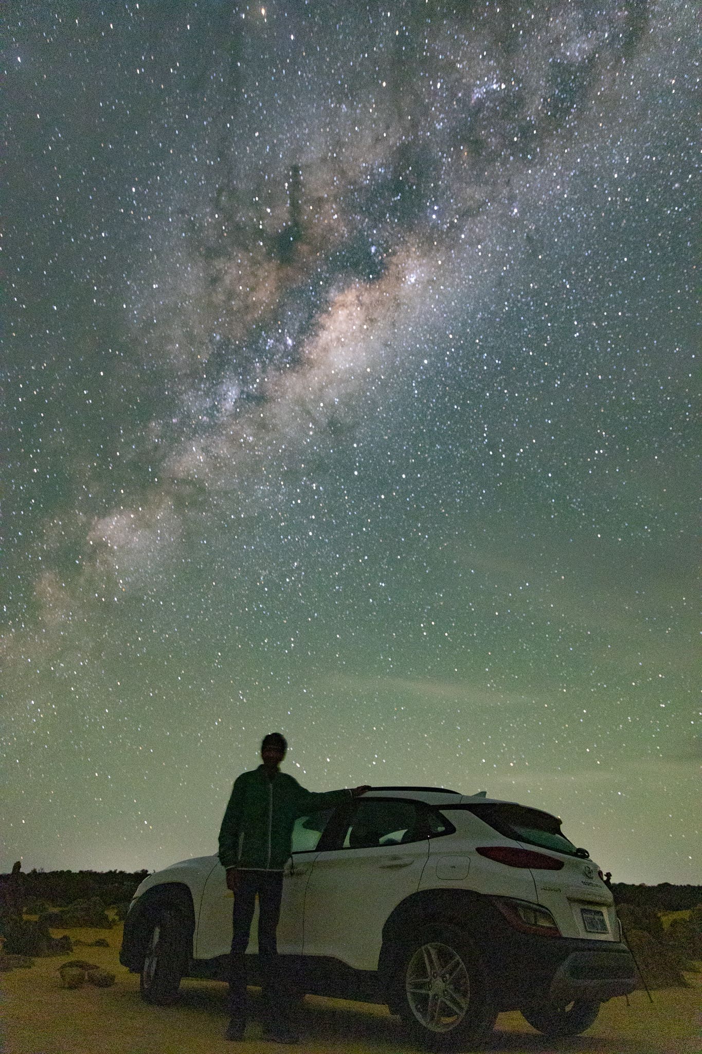 A person and a car silhouetted beneath a wide arch of the Milky Way in the Australian outback, with a green horizon glow.
