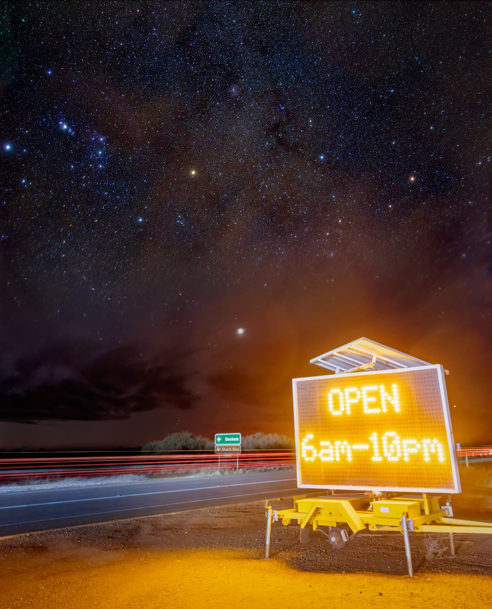 An illuminated Australian roadside sign glowing orange beneath a dense starfield and faint Milky Way band.