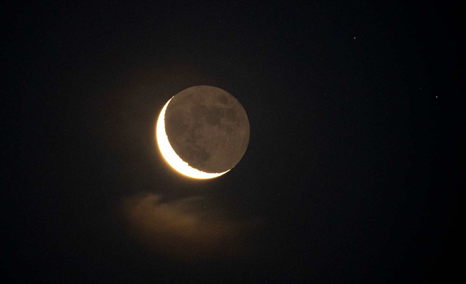 A thin crescent moon with its dark side faintly visible, glowing softly from earthshine against a twilight sky.