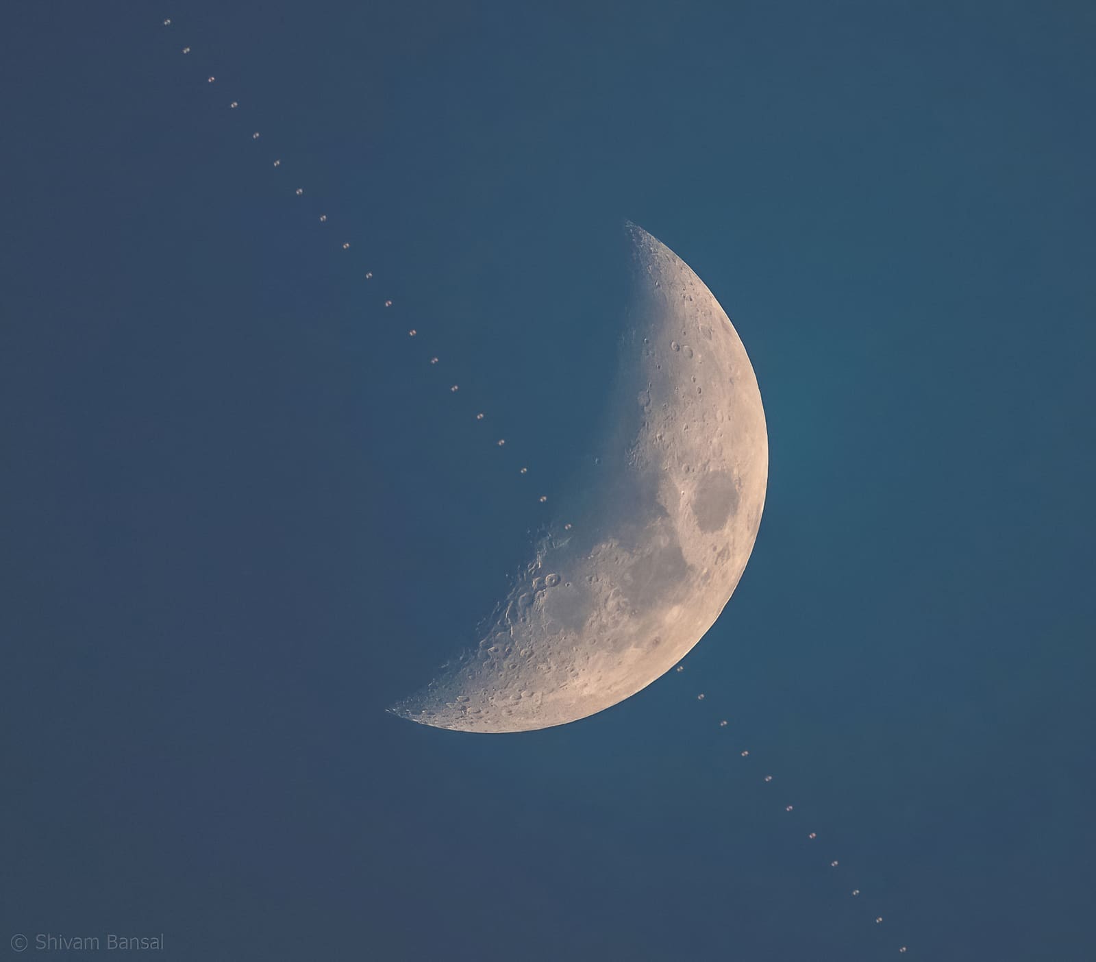 A crescent moon against a blue twilight sky with a thin diagonal streak crossing it, marking the transit of the International Space Station.