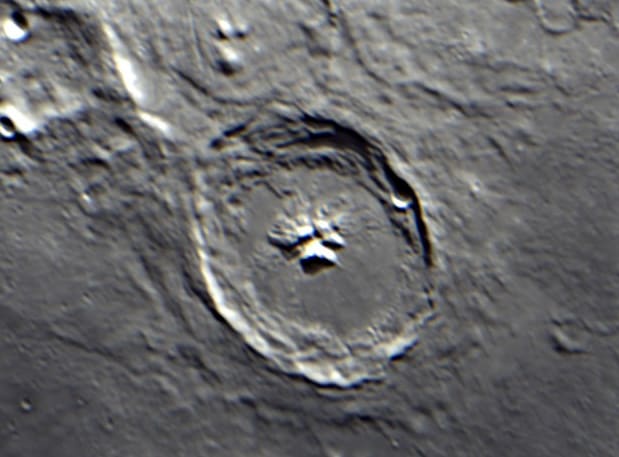A lunar crater with a tall central peak casting a long shadow across its floor, terraced walls visible.