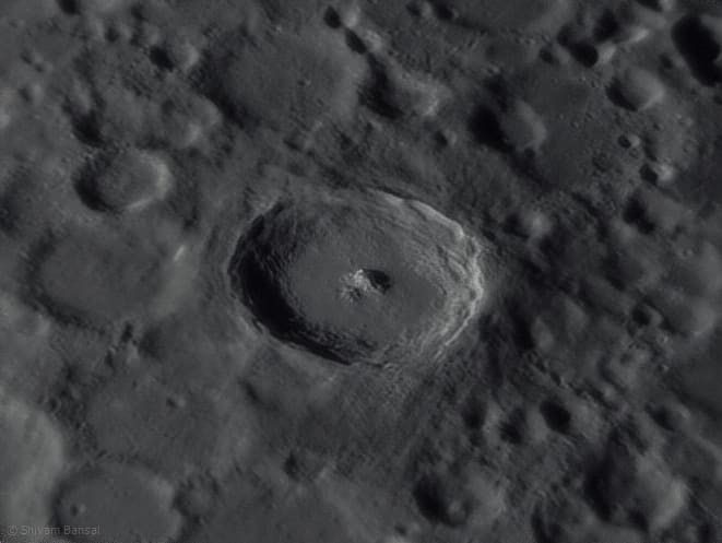 A close-up of a young lunar crater with a terraced rim and a central peak casting a shadow.