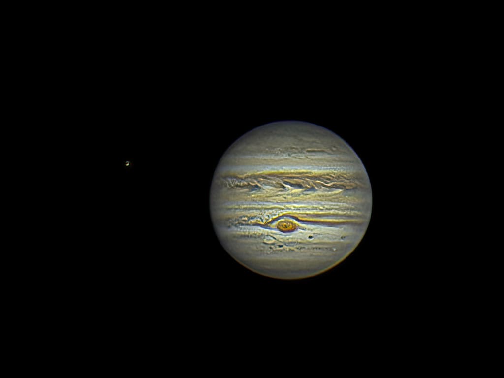 Detailed telescopic image of Jupiter showing banded clouds and the Great Red Spot, with its moon Io visible as a small dot to the left against black space.