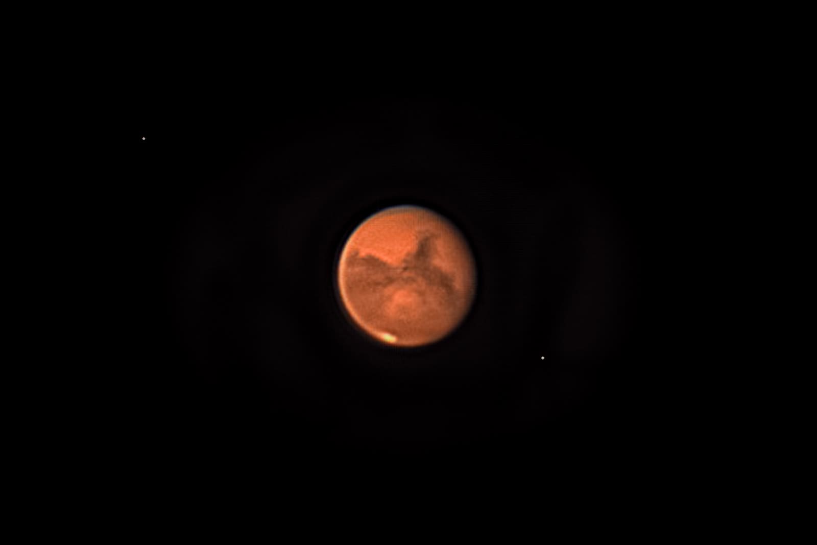 Telescopic image of Mars showing dark surface markings on its orange-red disc, with its two small moons visible as faint points of light nearby against black space.