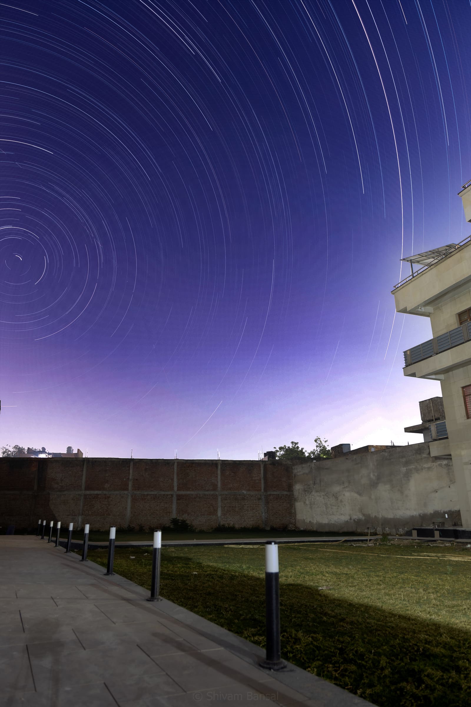 Long-exposure night photo of circular star trails over a courtyard with a walkway, lamp posts, a brick boundary wall, and a building to the right.