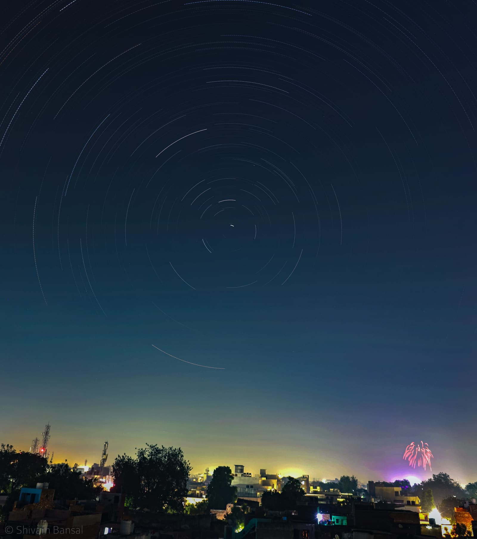 Circular star trail pattern in the night sky above a city skyline with illuminated buildings and a colorful firework burst near the horizon.