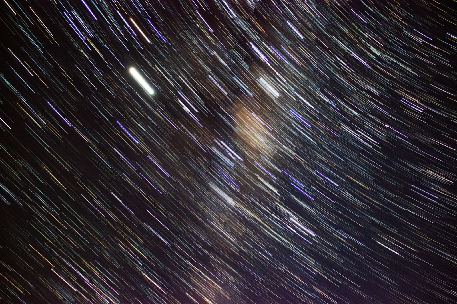 Diagonal colorful star trails crossing a dark sky with a diffuse pale glow of the Milky Way core visible in the center.