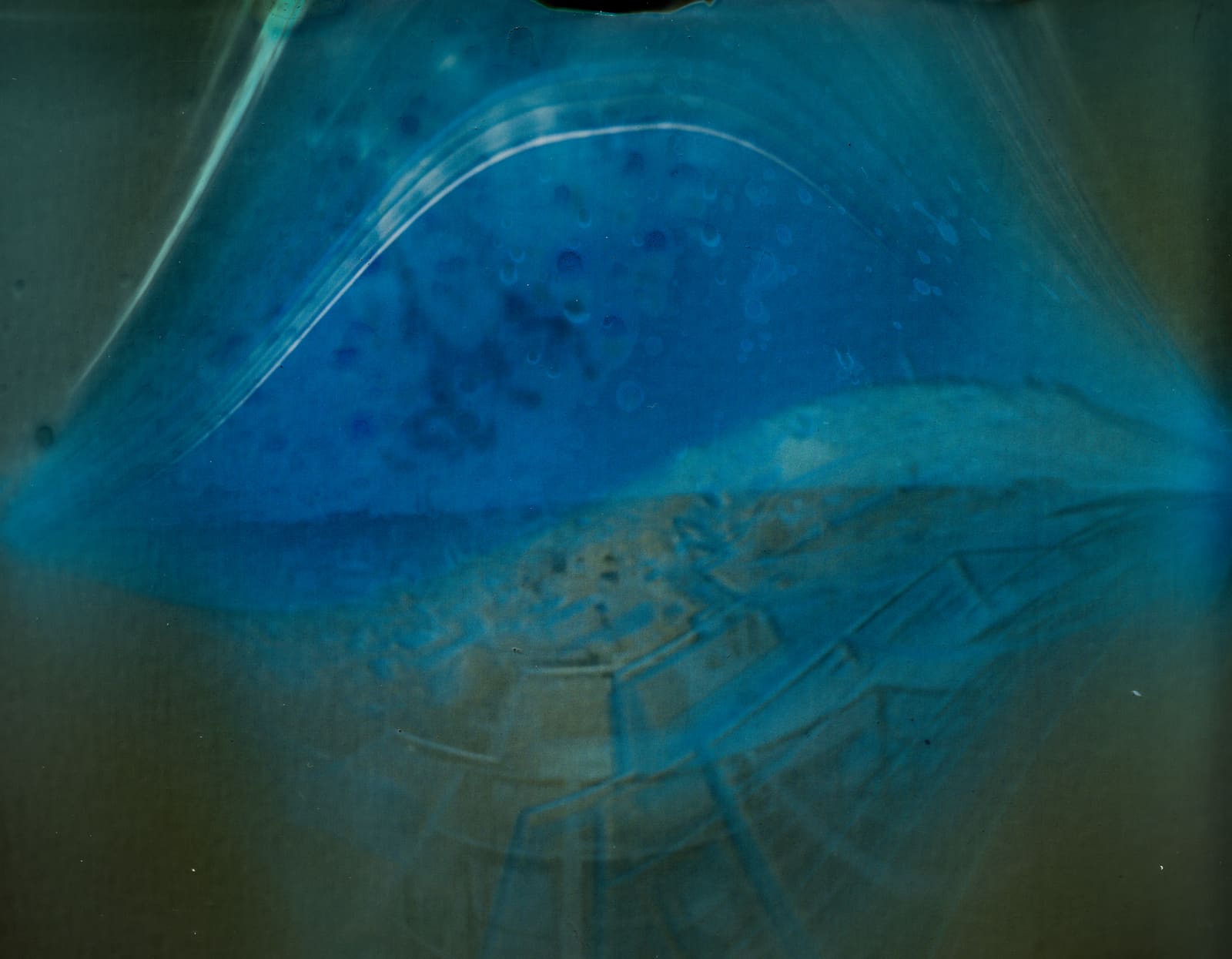Cyanotype-toned pinhole solargraph showing one prominent curved sun-trail arc over a dim silhouetted rooftop and railing, recorded over several months.