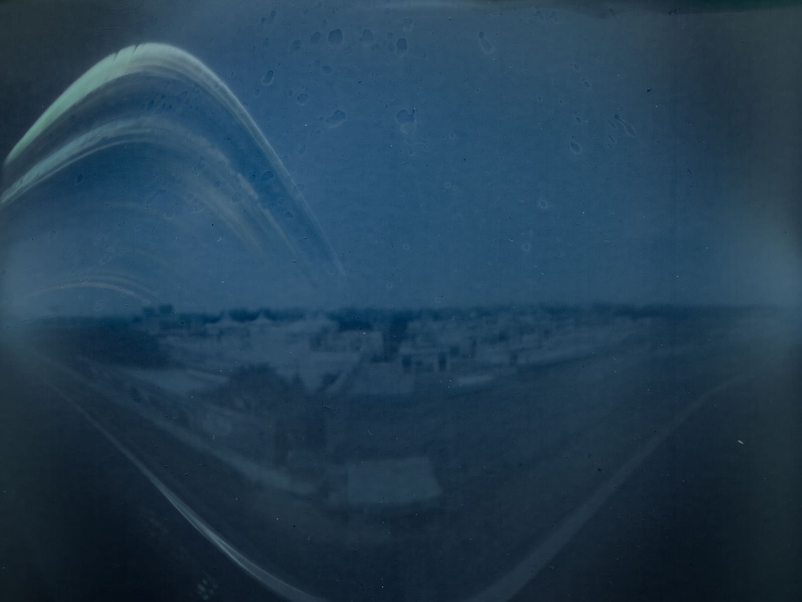 Cyanotype-toned pinhole solargraph showing multiple curved white sun-trail arcs over a faint silhouetted skyline, recorded over several months.