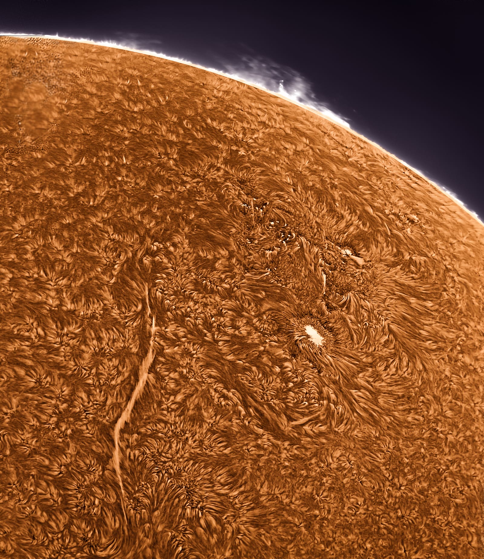 Close-up H-alpha image of the sun's chromosphere showing a long dark filament and a bright active region against mottled orange surface texture, with the limb visible in the upper corner.