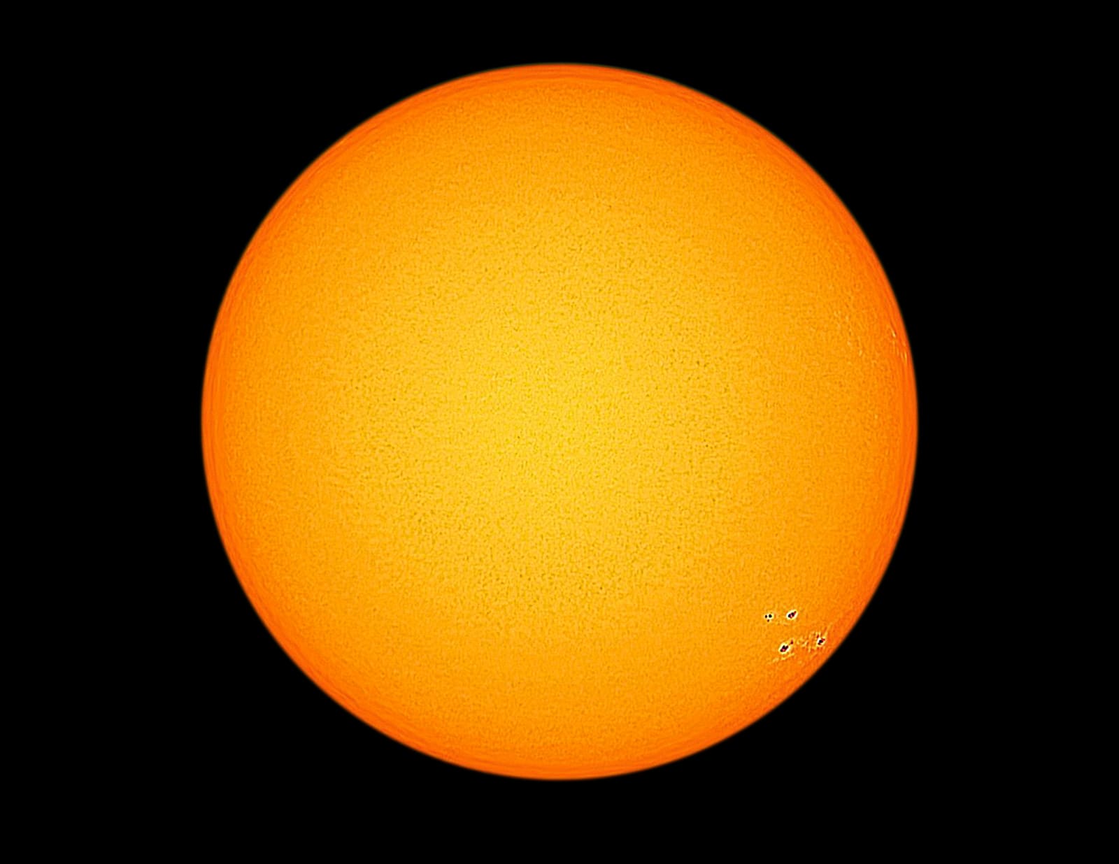 White-light photograph of the sun as a smooth orange disc with a small group of dark sunspots near the lower right, set against black space.